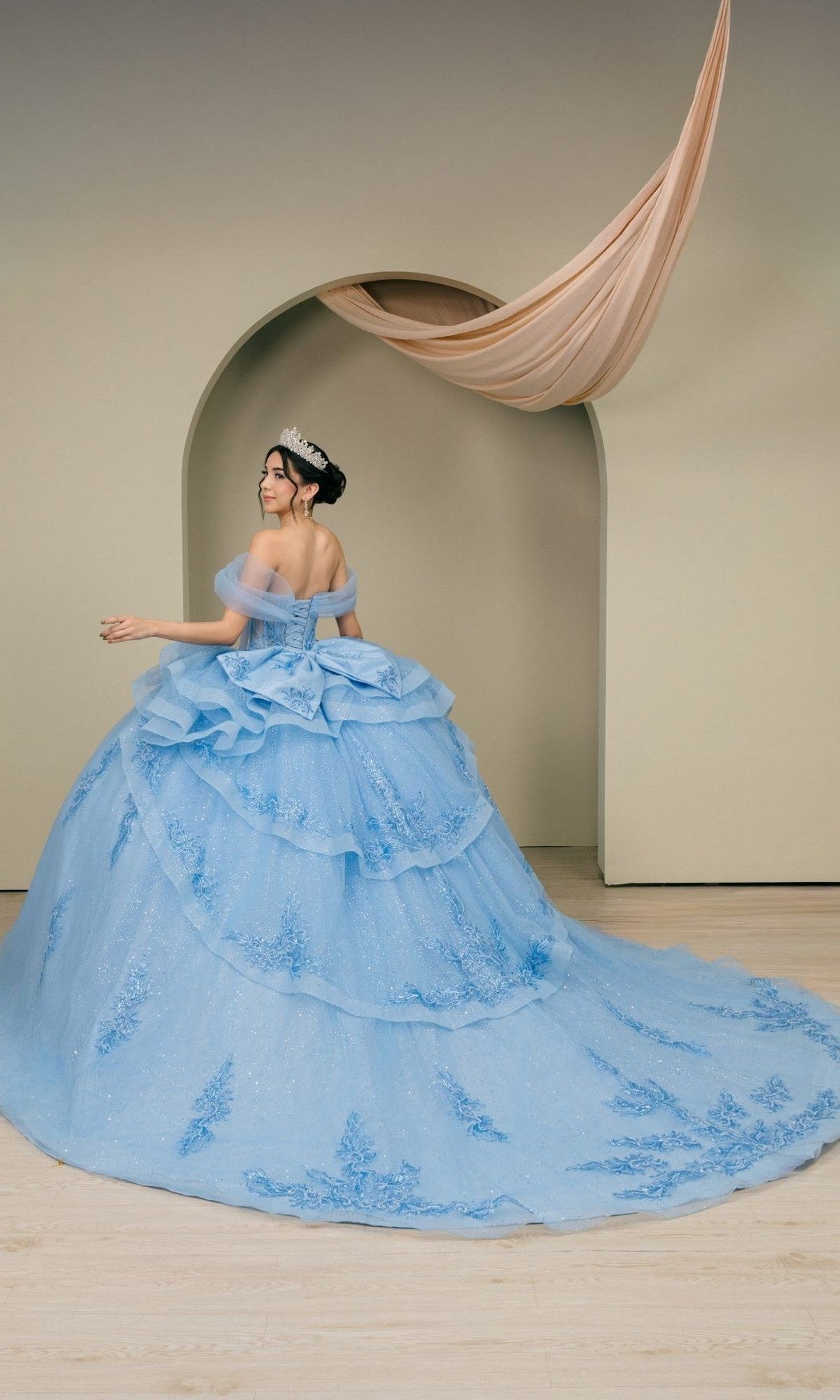 A person wearing the Quinceanera Dress 8520JQ By Cinderella Couture—an ornate light blue ball gown featuring a sparkling corset bodice and a tiara—stands in front of a beige arched doorway with fabric elegantly draped overhead.